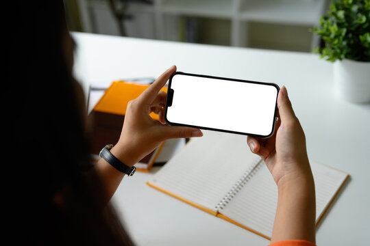 Hands holding smartphone with blank white screen in landscape orientation over a workspace