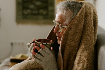Senior Caucasian woman sitting wrapped in blanket holding mug with both hands wearing glasses and...