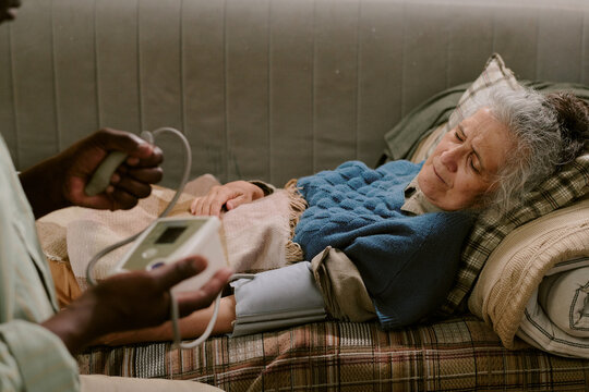 Senior Caucasian woman lying on couch with eyes closed, receiving blood pressure check from Black man using medical device, elderly couple experiencing health monitoring at home
