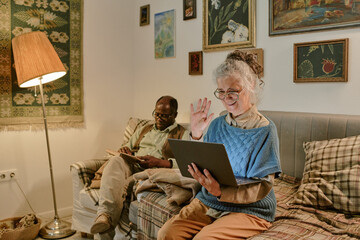 Senior Caucasian woman sitting on couch waving at laptop screen while senior Black man reading book beside her, both appearing engaged in separate activities in living room setting