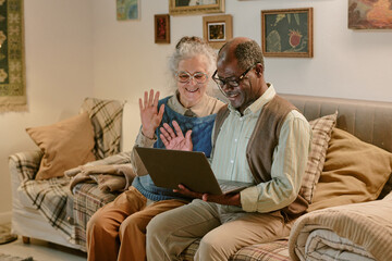 Senior Caucasian woman and senior Black man sitting together on couch waving at laptop screen,...
