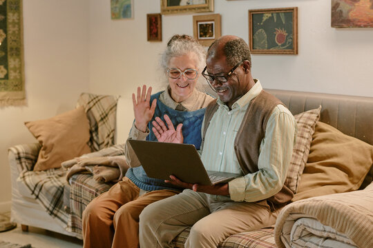 Senior Caucasian woman and senior Black man sitting together on couch waving at laptop screen, smiling and engaging in video call, framed by cozy home setting with wall art in background