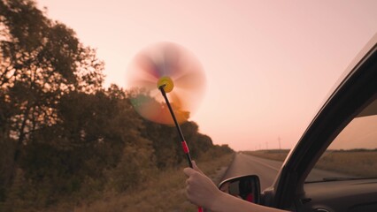 free girl waving a pinwheel out of the car window in wind, travel adventure road at pink sunset,...
