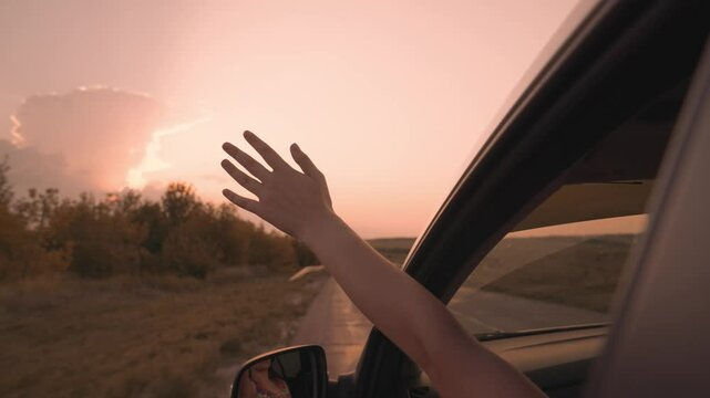 Free girl stretches out her hand from car in a pink sunset, catching wind with her hand while by transport on road, happy family, going on dream adventure vacation, having fun on road trip.