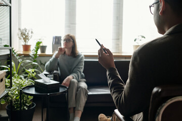 Woman sitting on sofa talking with man holding pen during therapy session in office setting with...