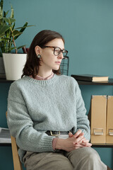 Woman sitting with hands clasped, wearing glasses, looking away thoughtfully during therapy...