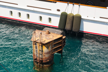 A luxury white yacht with protective fenders contrasts with an old, rusty mooring structure in the...