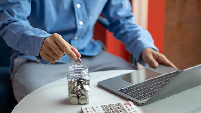 Person saving coins in a jar while managing finances on a laptop. Symbol of financial planning, budgeting, money saving, and smart personal expense management at home or office.