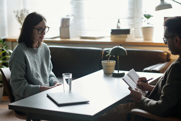Woman sitting on sofa talking to man therapist taking notes during therapy session in office...