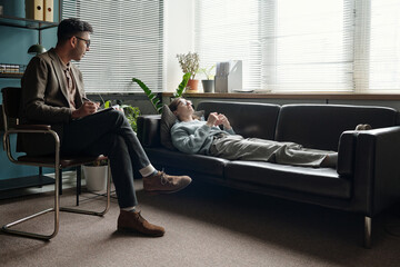 Woman lying on sofa talking to man therapist sitting with notebook listening attentively during...