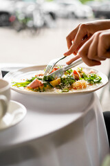 Close-up of a fresh green salad with tomatoes and cheese being eaten with a fork and knife at an outdoor table, suggesting healthy eating and a casual dining experience.