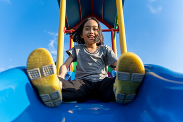 A little Asian girl playing at playground slide looking directly at the camera with a wide, joyful...