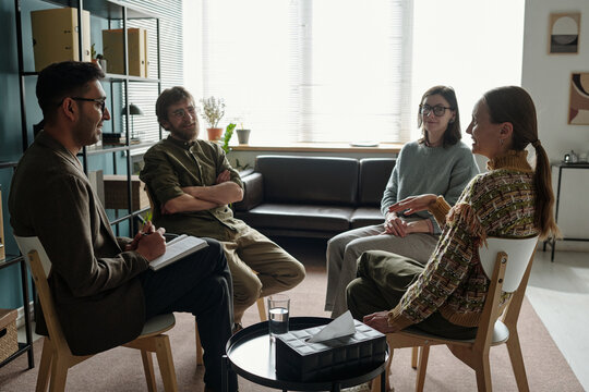 Group of multiethnic men and women participating in therapy session, sitting in circle, engaging in discussion with therapist holding clipboard