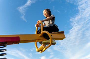 A happy young Asian girl on a yellow seesaw at a playground. Happiness, freedom, childhood fun, and outdoor activity concept.