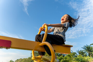A happy young Asian girl on a yellow seesaw at a playground. Happiness, freedom, childhood fun, and outdoor activity concept.