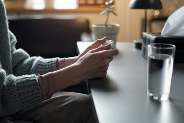 Woman sitting at table holding tissue with both hands during therapy session, glass of water and...