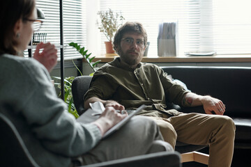Man sitting on sofa engaging in therapy session with woman therapist holding clipboard, both facing each other in professional office setting