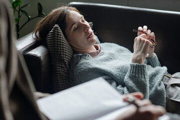 Woman lying on couch holding eyeglasses during therapy session, therapist with notepad listening...