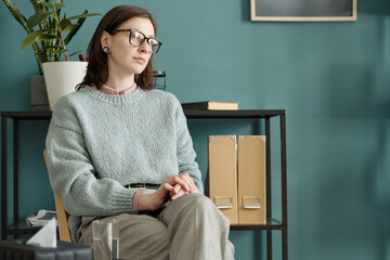 Woman sitting with hands clasped, wearing glasses, looking away thoughtfully during therapy session...