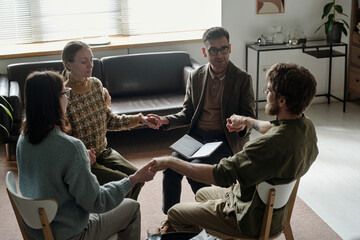 Group of and Black people sitting in therapy session holding hands in circle, male therapist with...