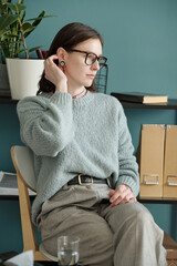 Woman sitting in therapy session touching ear, wearing eyeglasses, looking away from camera,...