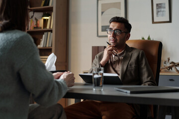 Man sitting at desk holding pen listening attentively to woman during therapy session in office setting with tablet and glass of water visible