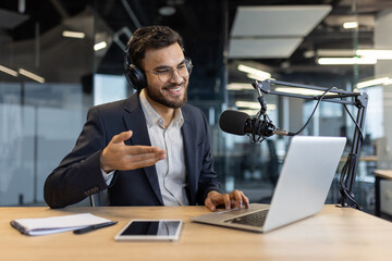 A businessman in a suit smiles while podcasting, wearing headphones, using a laptop, and speaking...