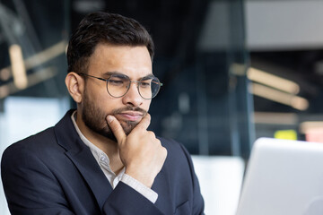 A thoughtful businessman with glasses considers his options while working on his laptop in a modern...