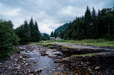 Fototapeta premium A serene mountain landscape with rustic wooden cabins nestled among dense pine trees. A small stream flows through the foreground, and misty hills rise in the background under a cloudy sky.