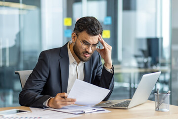 A concerned businessman reviews documents in his office, displaying stress and concentration as he...