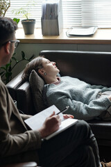 Woman lying on couch during therapy session with man taking notes, both seated in professional...