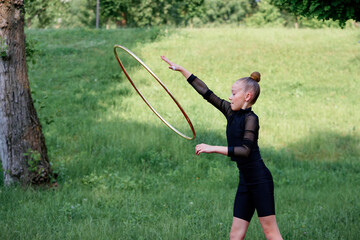 Young gymnast girl wearing black exercise apparel performs a rhythmic gymnastics routine with hula hoop on green lawn under trees. Young Girl Practicing Rhythmic Gymnastics Outdoors with Hula Hoop