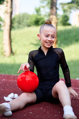 Girl in a black sporty outfit practicing rhythmic gymnastics on a sunny day with her red ball,...