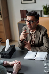 Man wearing glasses conducting therapy session, gesturing while speaking to unseen client, notebook...