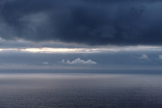 Dark storm clouds gather over a calm, gray ocean with a bright line of light on the horizon - Powered by Adobe
