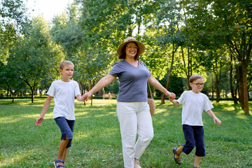 Joyful outdoor scene of a mother with her kids holding hands and walking surrounded by greenery in...