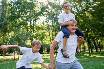 A happy family enjoying time together outdoors in a lush green park setting with trees. Father running with his sons in the park.