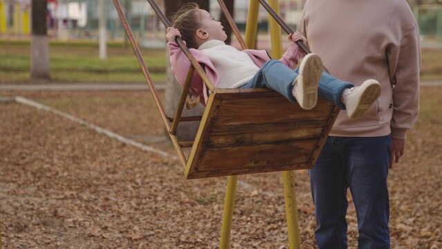 father shakes the little kid on playground swing, child dreams of flying, happy family, smiling laughing child in free flight, dad and daughter play game on street in park, girl cheerful emotions
