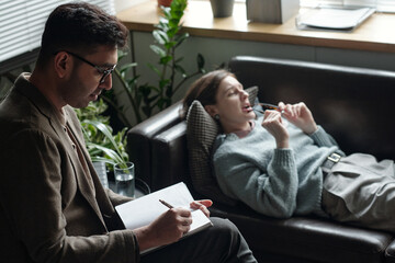 Woman lying on couch talking while man sitting nearby writing in notebook during therapy session in office setting