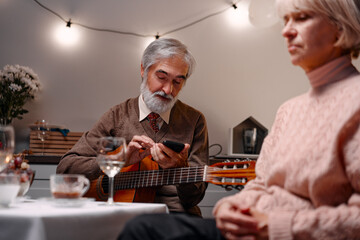 An elderly man uses his phone while holding a guitar, set against a warmly lit room with an...