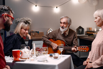 A family spends time together enjoying a cozy dinner while an elderly man plays guitar. The atmosphere is warm and intimate, showcasing smiles, food, and companionship in a well-lit dining room.