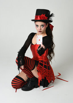 Burlesque circus performer, fantasy female model in halloween clown costume with top hat, red gothic corset holding  ace playing card. theatrical sitting pose, isolated studio background