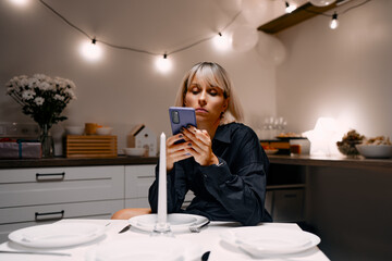 A woman occupies time reading her phone while seated at a dinner table set with plates and decorations in a cozy, warm-lit environment with decorative lighting and soft-colored furniture.