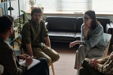 Group of and men and women participating in therapy session, sitting in circle, engaging in discussion, therapist holding clipboard, natural light from window