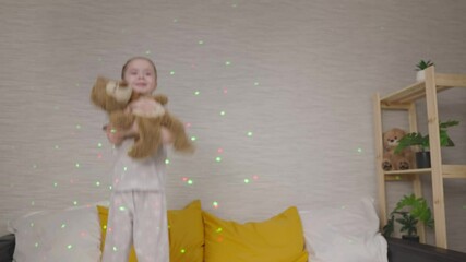 little child is jumping with a teddy bear in his hands on bed in bedroom, kid girl is playing with toy, cheerful life is dancing in bright multi-colored garlands of light, happy daughter is having fun - Powered by Adobe