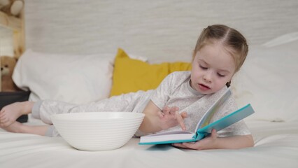 little child is reading book while lying in bed children's room, doing homework first grade,...