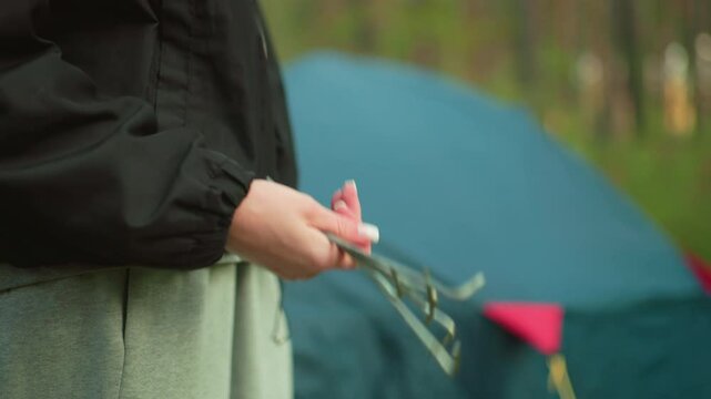 Close up of lady holding and twirling metal pegs outdoors with forest camping tent in background, wearing casual outfit, preparing for camping setup in nature with blurred greenery and gear in view