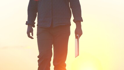a farmer in boots walks in the sunset on the road with a tablet, the use of modern gadgets in...