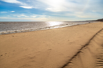 Diagonal view of Gulf of Finland coast with wind-shaped yellow sand dunes and melting ice remnants. Backlit pine forest and distant silhouettes under hazy blue sky. Komarovo, Russia. 