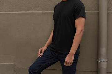Mid-adult man is standing against beige wall, flat, wearing black tee, jeans, ledge and drain pipe